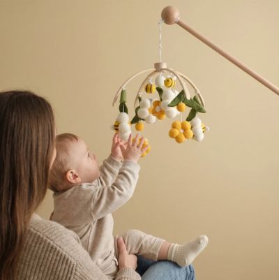Maman tenant son bebe qui regarde et tend les mains vers le mobile bebe en bois fleurs et abeilles